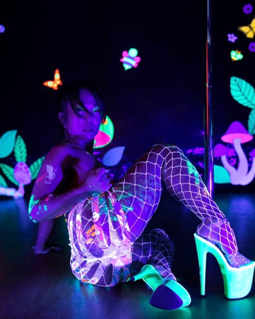 A pole dancer in heels takes a photo in a blacklight forest themed shoot