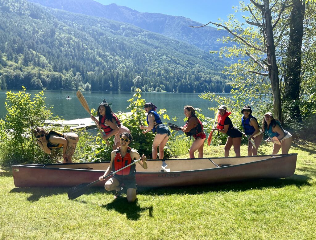A group of 8 people posing with a canoe next to a sunny lake.