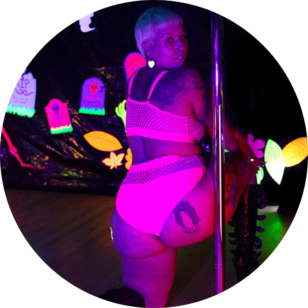 Katie is doing a lunge facing away from the camera and next to the pole. They are looking over their shoulder and illuminated in black light neon pink.