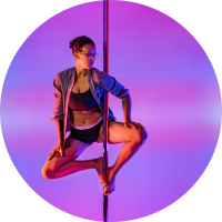 BIPOC person with short dark hair and glasses doing a pole sit variation. They are wearing a button down shirt, black sports bra, and black bottoms. There is a trans flag in the background.