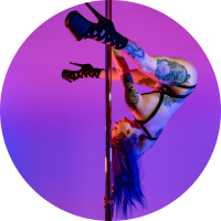 White femme person with purple hair and tattoos does an ayesha on the pole. They are wearing a black strappy one piece, black strappy heels, and are looking away from the camera. The background is the bisexual pride flag colors.