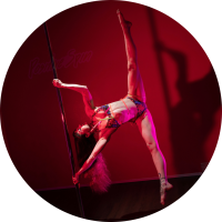 White femme person with long colorful hair doing an iron X on the pole. They are wearing a strappy pole set. The lighting is dark and they are against a pink wall and casting a shadow against it.