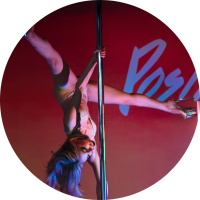 A white fem-presenting person does a true-grip ayesha in dark rainbow lighting on the pole. They have long hair and you can see part of the Positive Spin logo behind them.