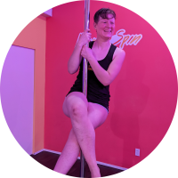 White non-binary person with short brown hair doing a pole sit facing the camera and smiling. They are having a blast on the pole!