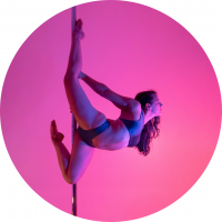 White femme person with long dark curly hair does a pole move called the lois lane. They are gazing off to the side and wearing a dark two piece pole set. The background is a pastel pink.