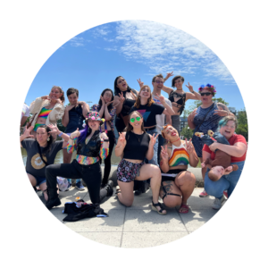 A group of pole dancers wear pride themed outfits and smile outside together